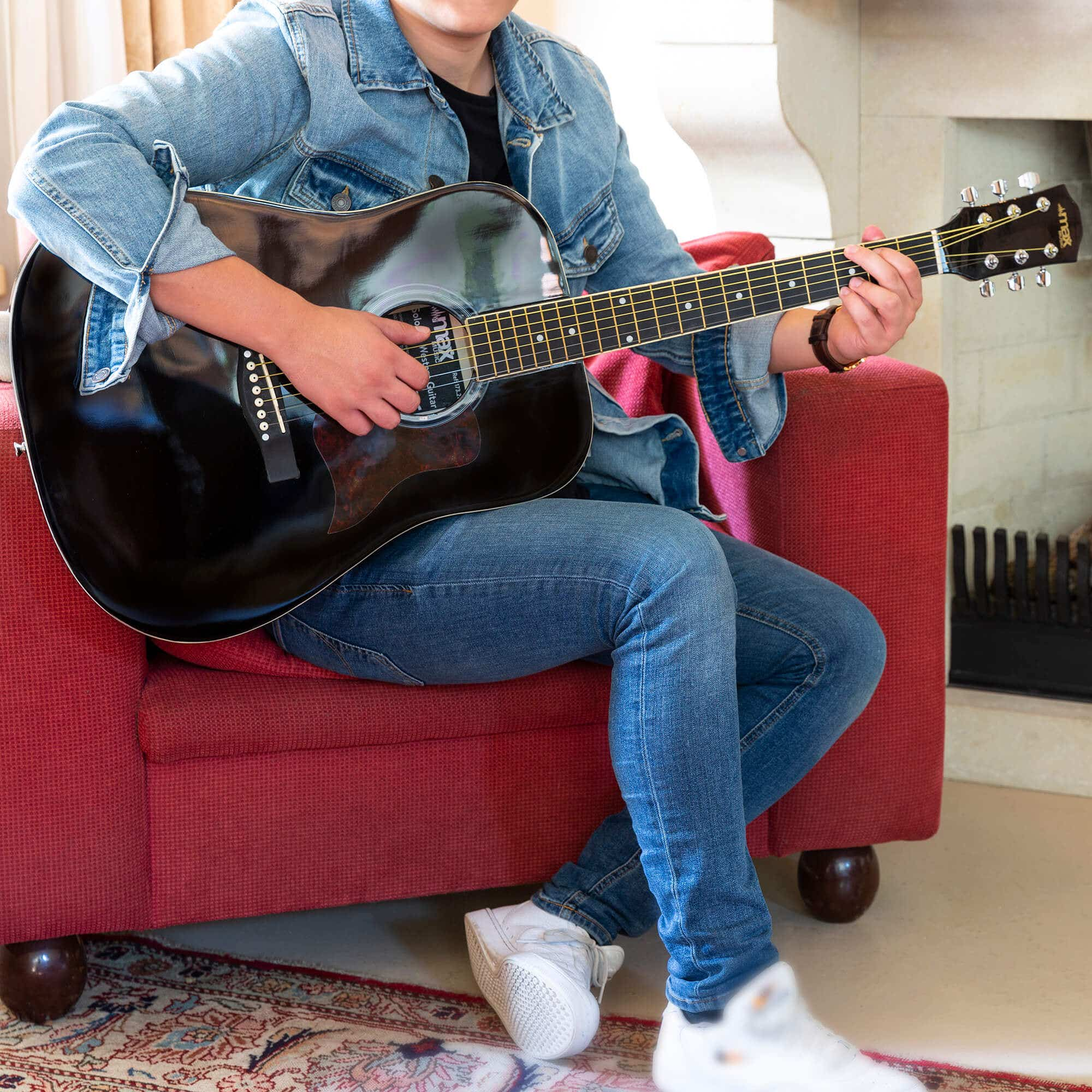Persoon speelt op een zwarte gitaar op een rode bank. Jeans en spijkerjasje. Interieur.