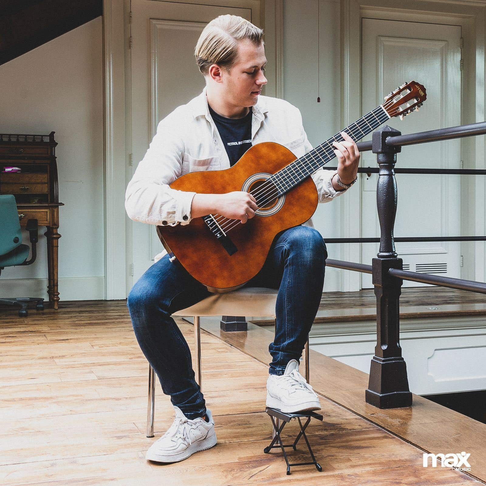 Mann spielt Akustikgitarre. Sitzt in einem Raum mit Holzboden. Weiße Turnschuhe.