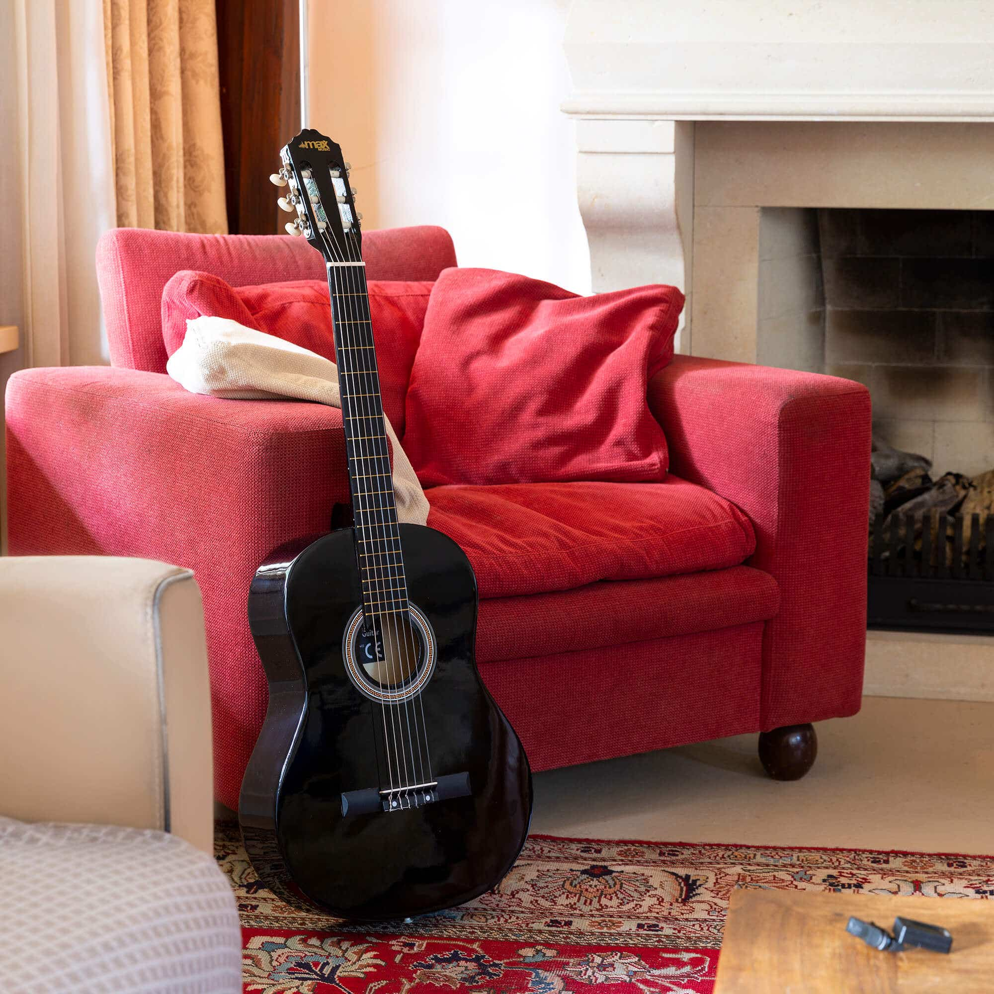 Guitarra negra apoyada en sillón rojo. Chimenea al fondo. Habitación con alfombra y mesa.