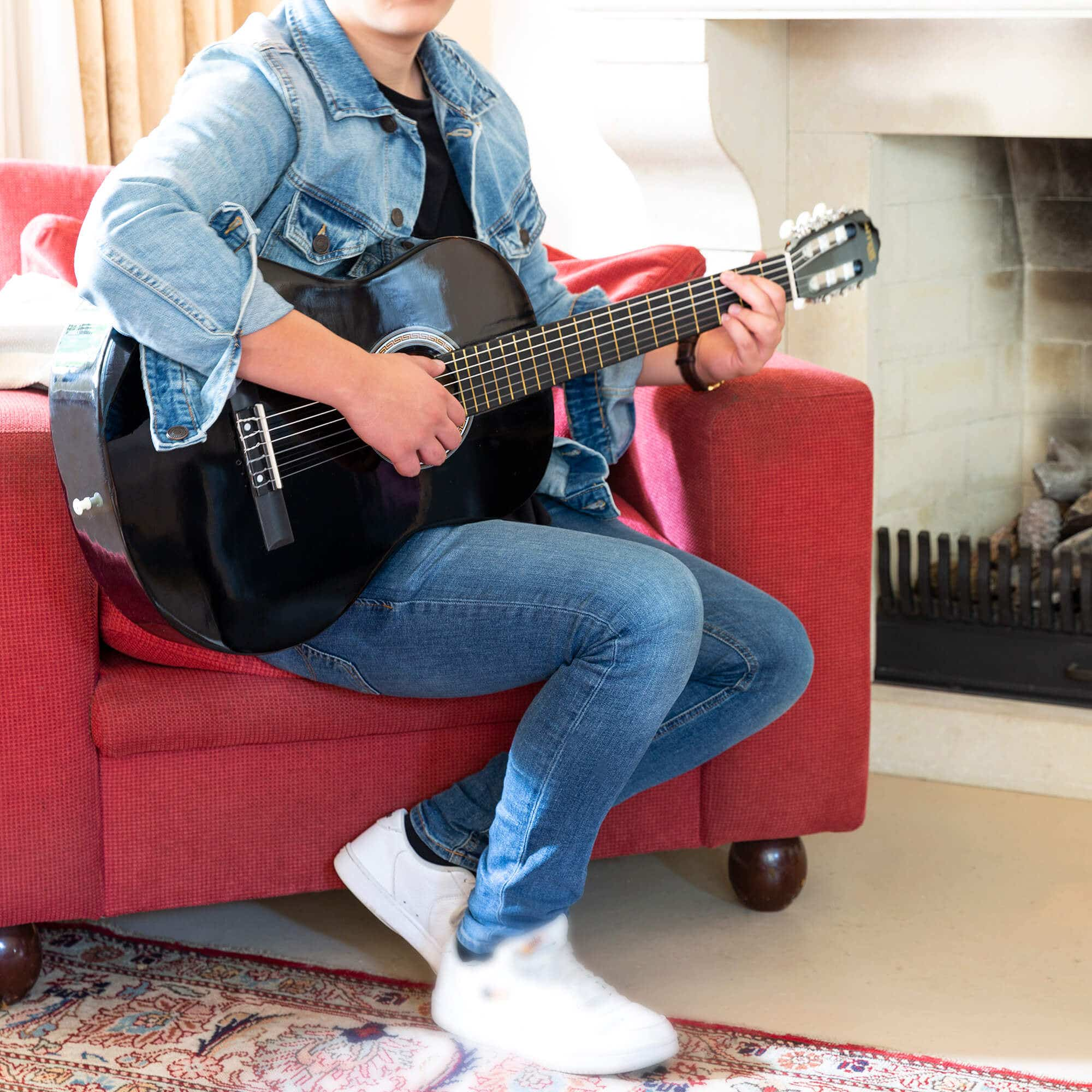 Persona toca una guitarra negra, sentada en un sofá rojo cerca de una chimenea.