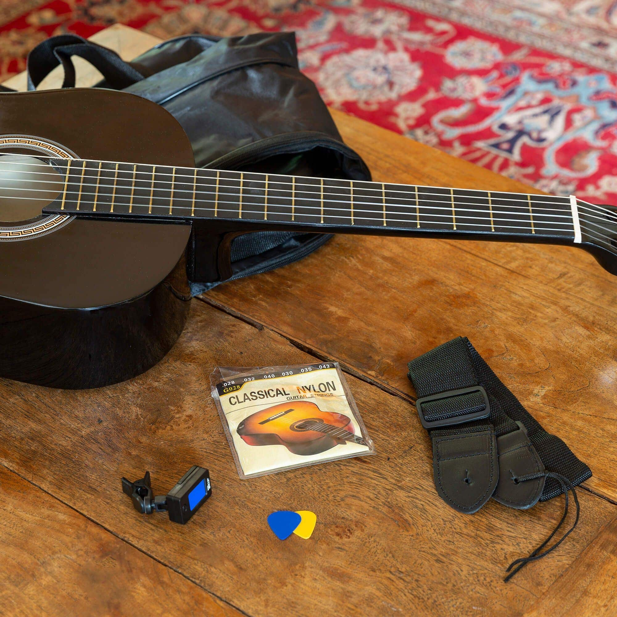 Guitarra, cuerdas, afinador, púas en mesa de madera. Bolsa negra al fondo. Alfombra roja.