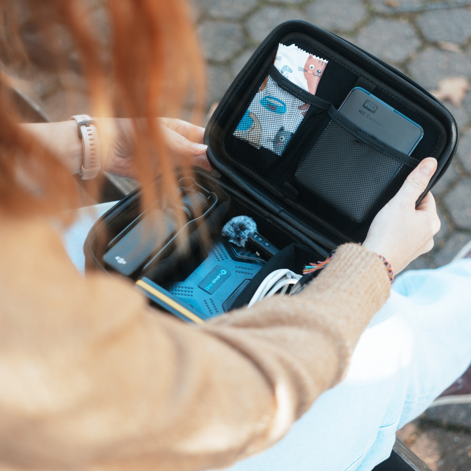 Eine Person mit ingwerfarbenem Haar hält eine offene schwarze Tasche, die ein Mikrofon und andere elektronische Geräte zeigt.