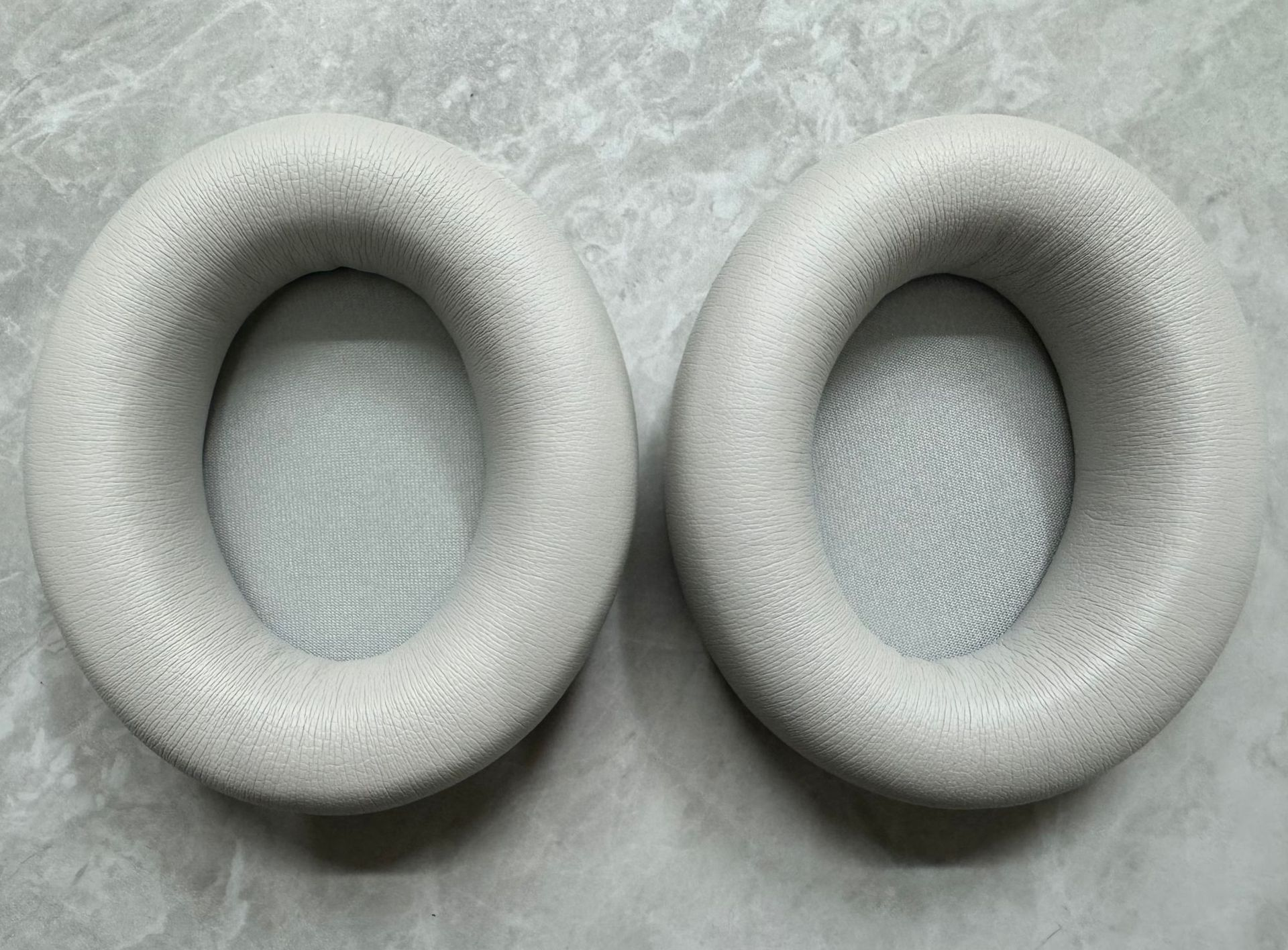 Dwie jasnoszare nausznice do słuchawek, okrągłe z centralnym otworem.
