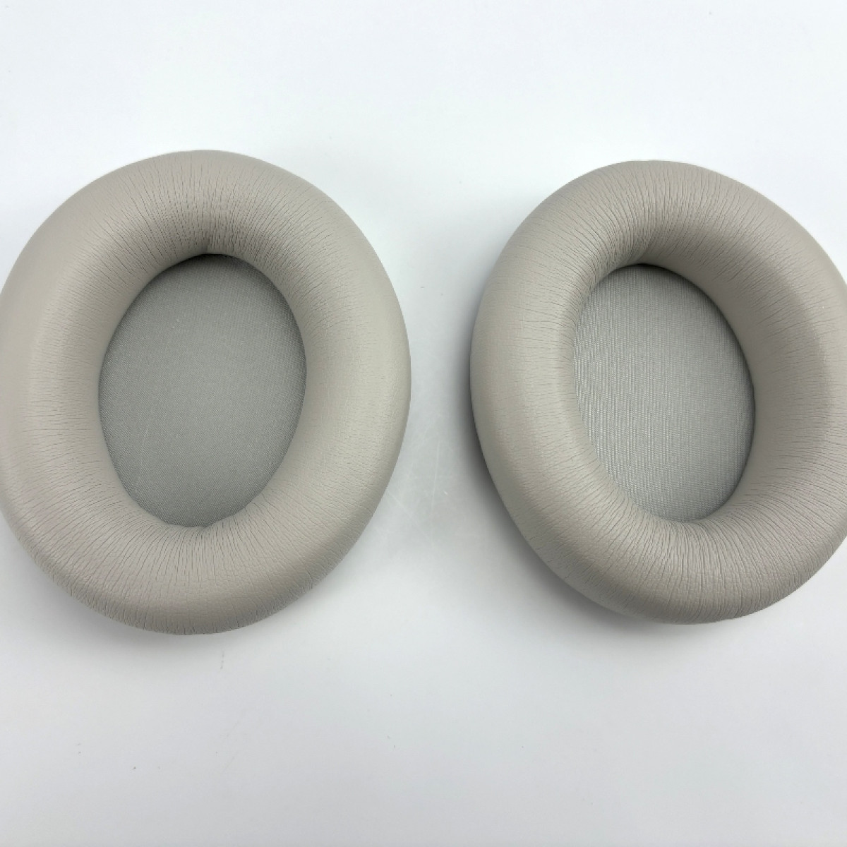 Dwie jasnoszare nausznice do słuchawek, okrągłe z centralnym otworem.