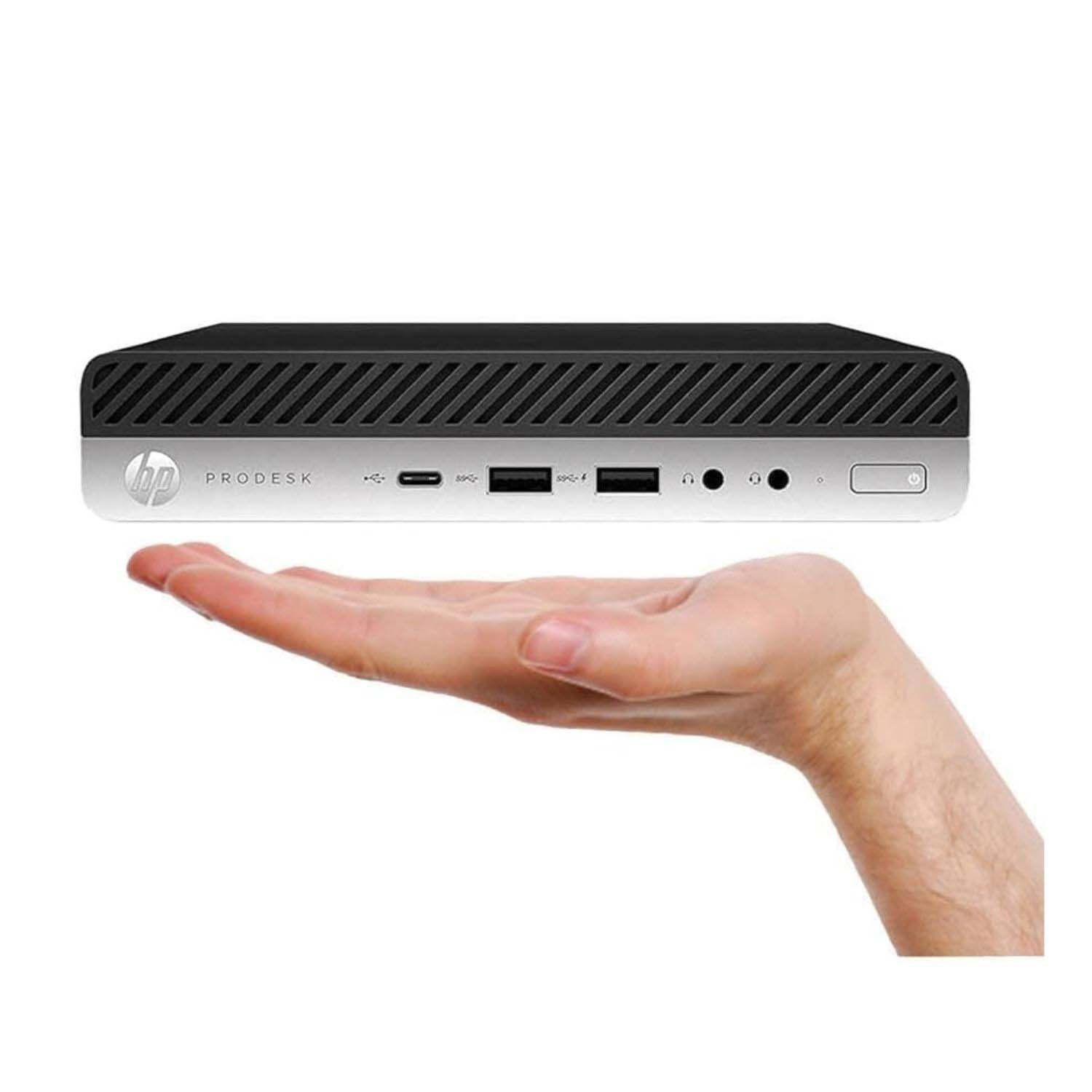 Mini PC negro y plateado sostenido por una mano. Tiene puertos. Fondo blanco.