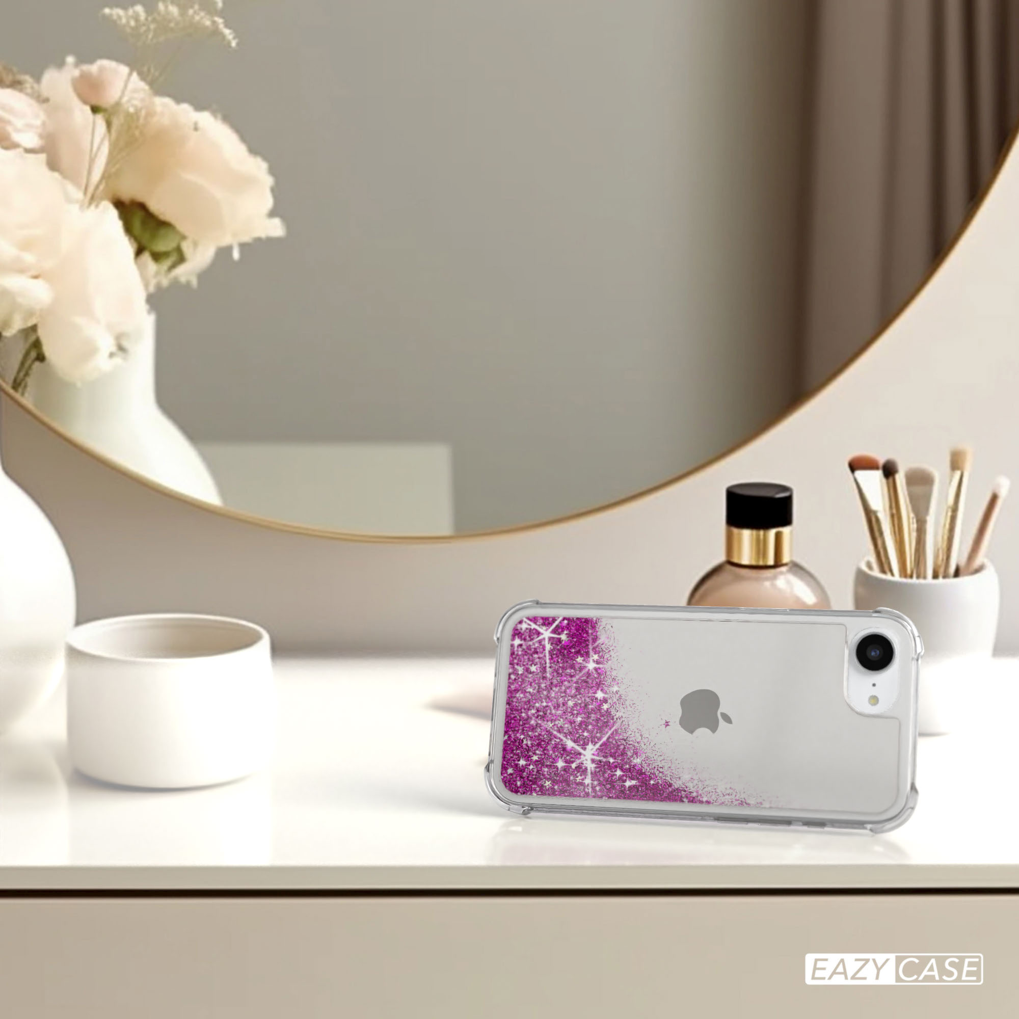 Telefon mit pinkfarbener Glitzerhülle auf einem Schminktisch neben Spiegel und Blumen.