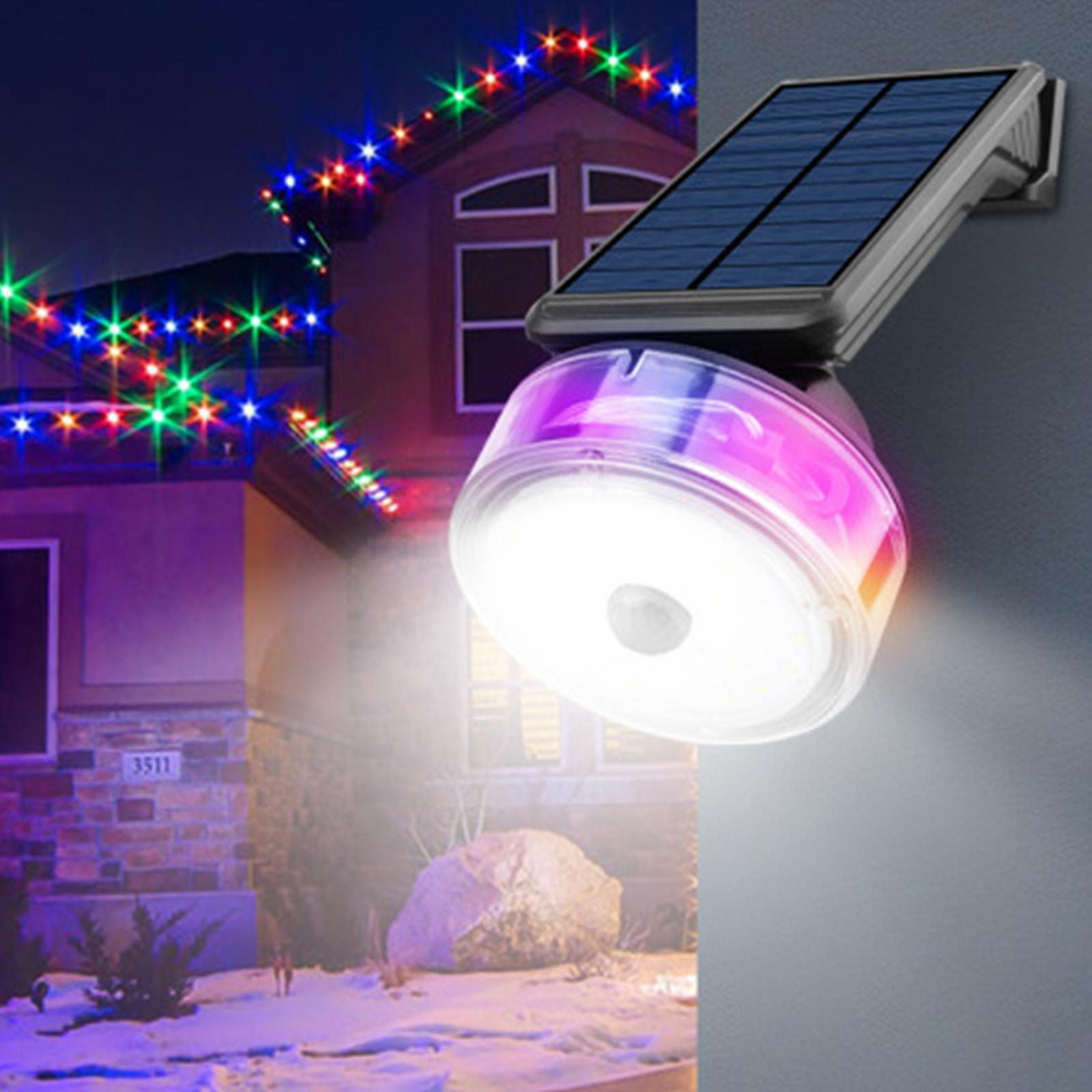 Solarbetriebene Leuchte an einer Wand. Haus mit Weihnachtsbeleuchtung im Hintergrund. Licht emittiert Farben.