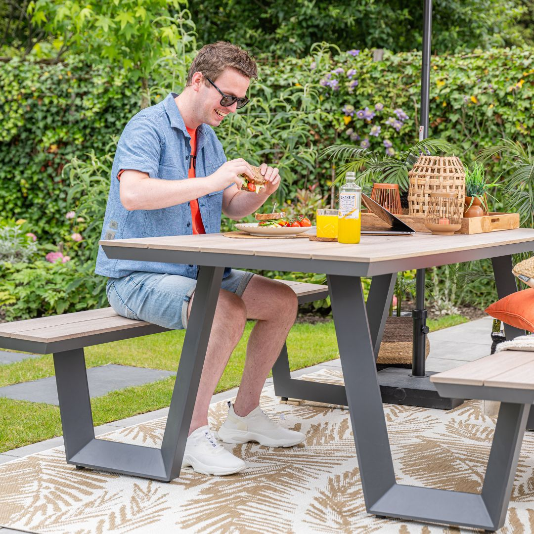 Man zit aan een tafel. Tafel met eten en drinken. Groene struiken op de achtergrond.