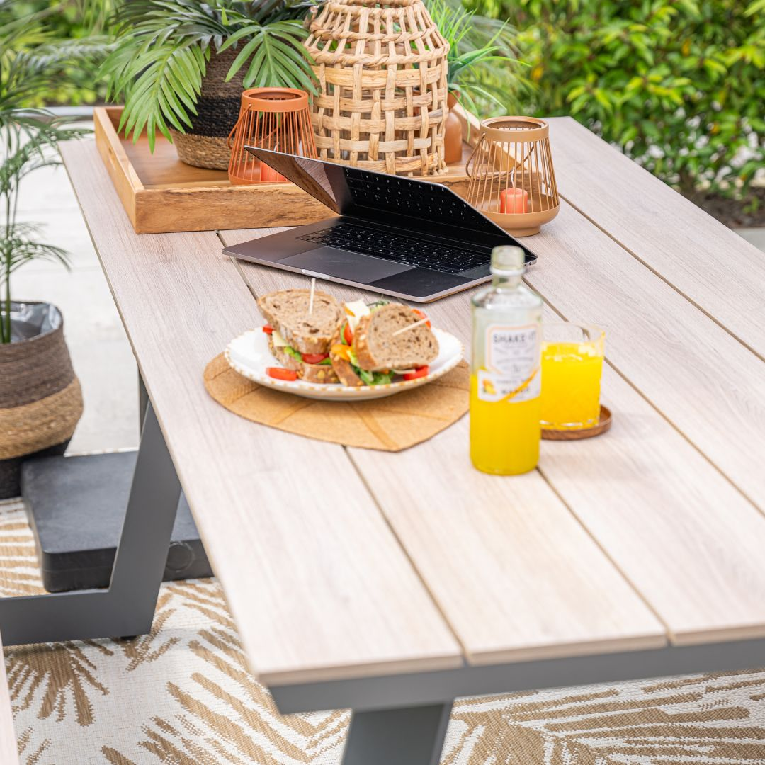 Buitentafel met eten, laptop, planten en sinaasappelsap, met een kleed met bladeren.
