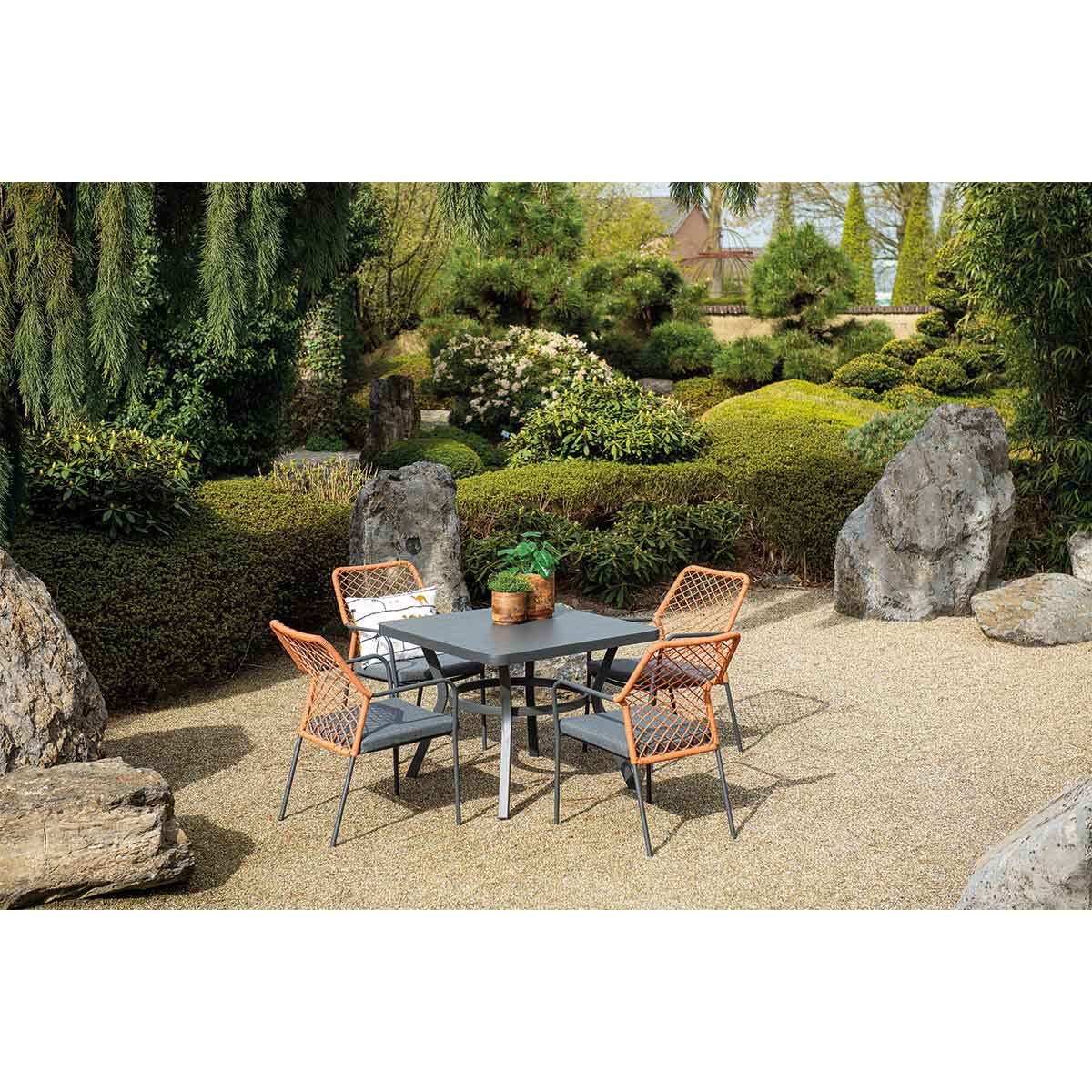 Eetset voor buiten: donkergrijze vierkante tafel, vier oranje stoelen, kiezelgrond, groene planten.
