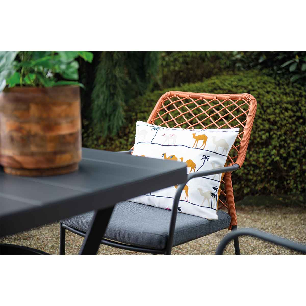 Buitenstoel met oranje geweven rug, kameelkussen en uitzicht op groene planten en een tafel.