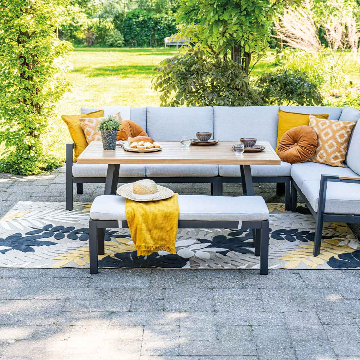 Outdoor patio setting with table, bench, couch, rug, pillows, and decor. Bright, sunny day.