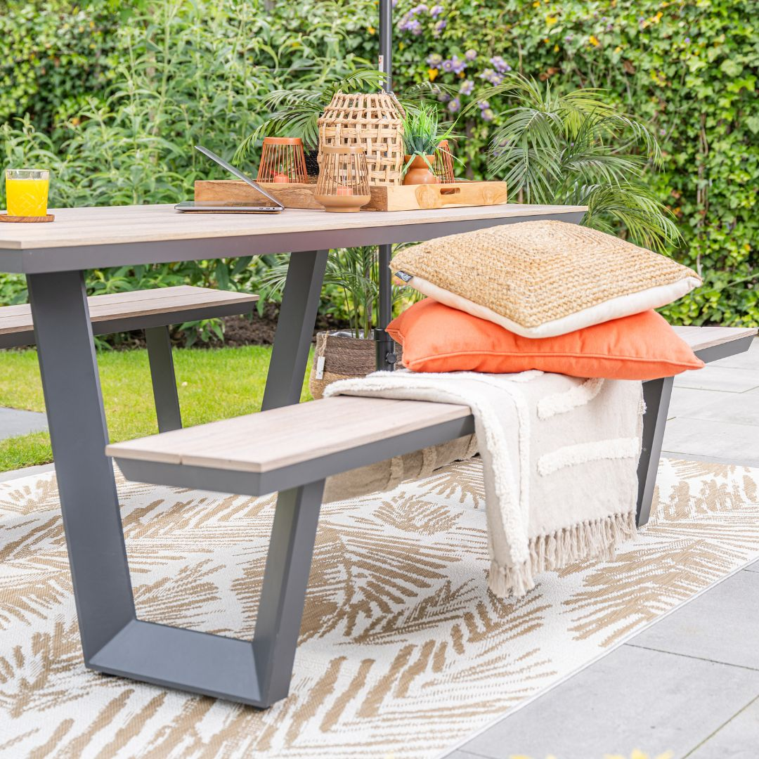 Buitentafel met bank, kussens, kleed en planten. De scène is helder.