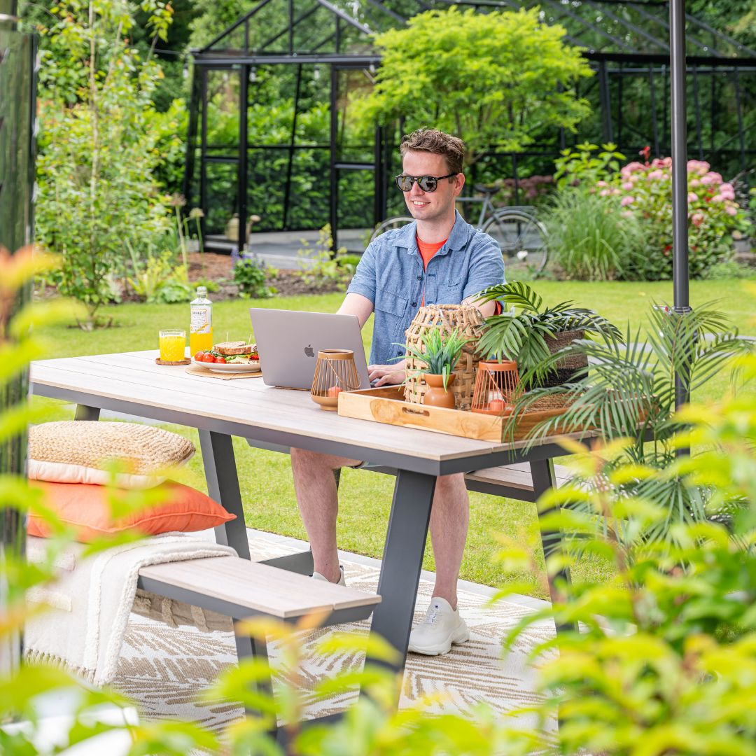 Man die op een laptop werkt aan een buitentafel met eten en planten, wazige achtergrond.