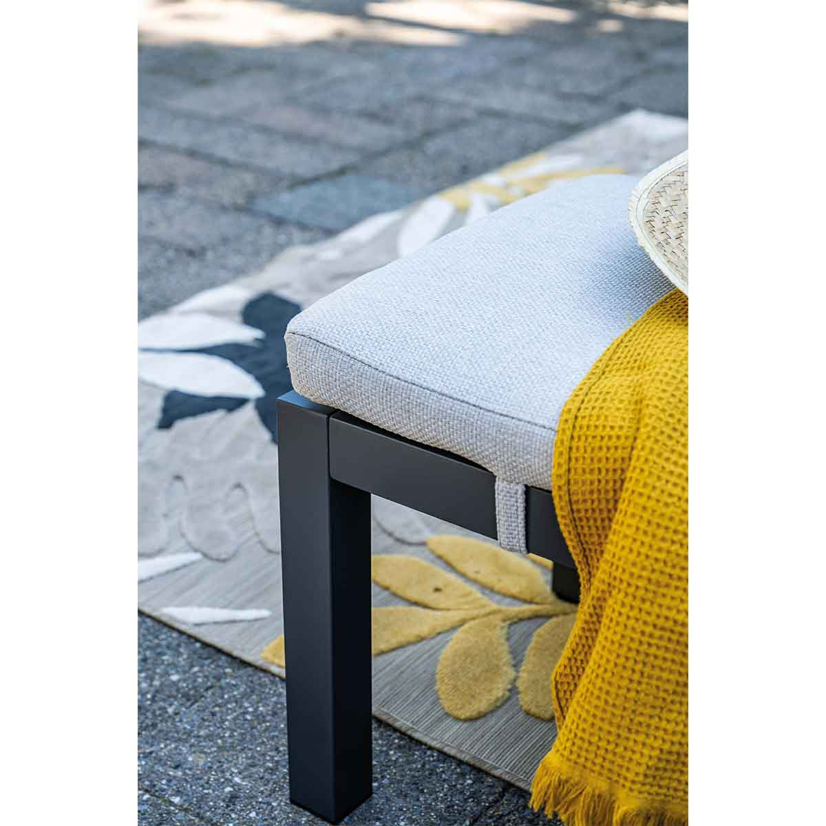 Bench with a gray cushion, yellow blanket, and straw hat. A floral rug is under it.