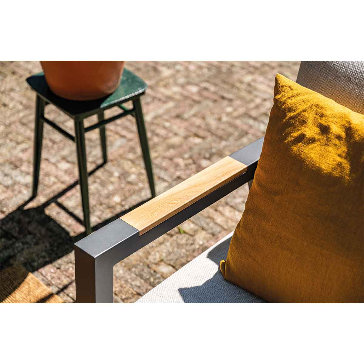 Stoelarm detail: houten armleuning op grijs frame, gele kussen, kleine groene kruk.