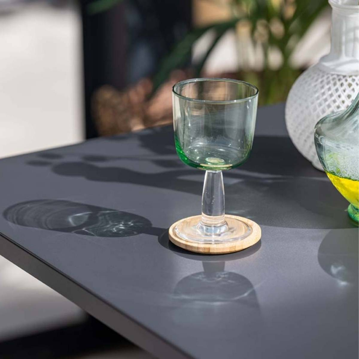 Een groen glas op een houten onderzetter, op een donkergrijze tafel. Ander glas en een vaas op de tafel.