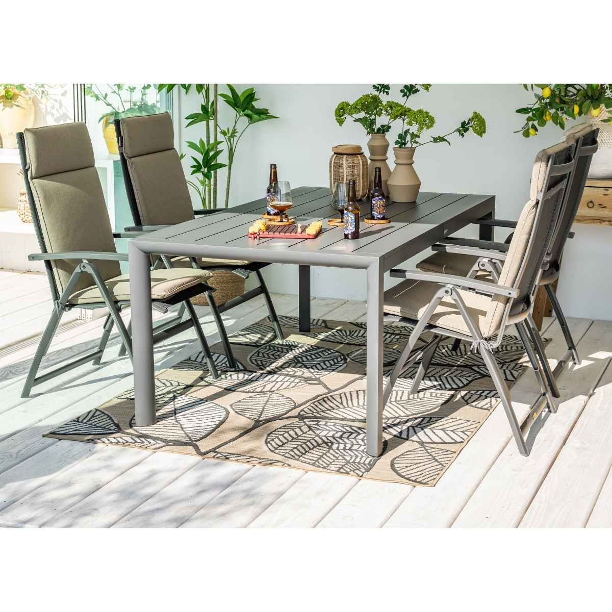 Outdoor dining set: gray table, chairs, rug. Bottles on table. Plants in background.