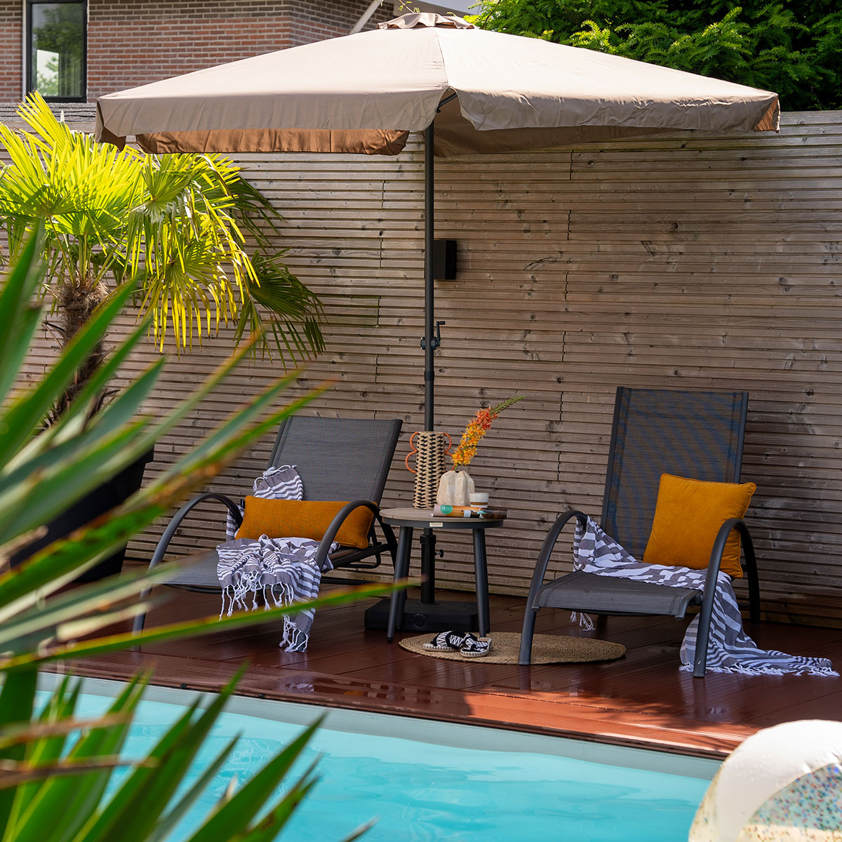 Two chairs near a pool, a table with a parasol, and a palm tree.