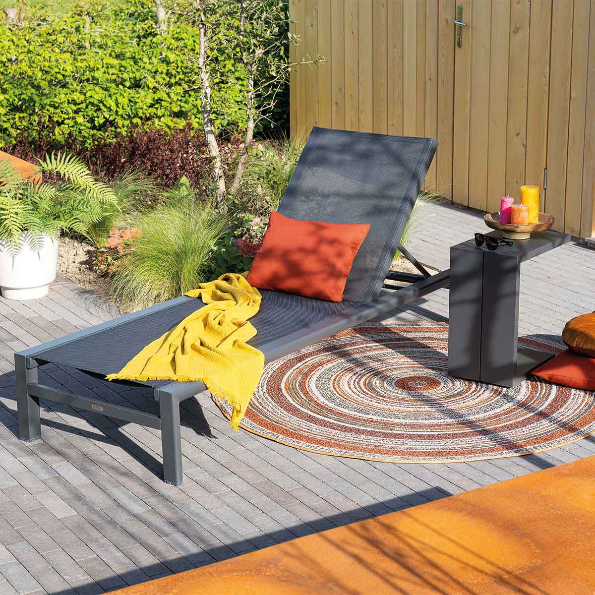 A chaise lounge with cushion, blanket, and side table in a garden setting.