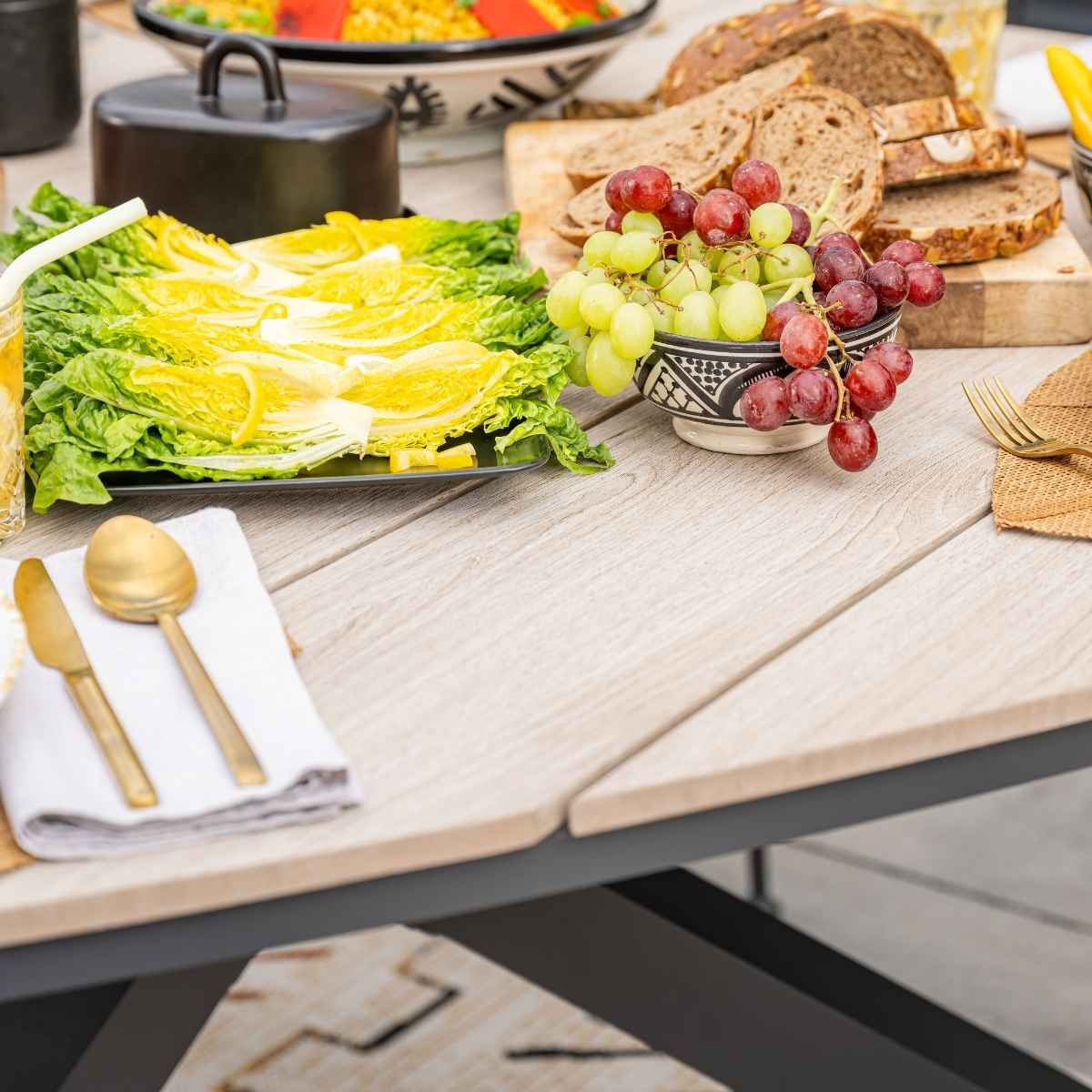 Table with food: green lettuce, grapes in a bowl, bread, gold utensils, and a black covered dish.