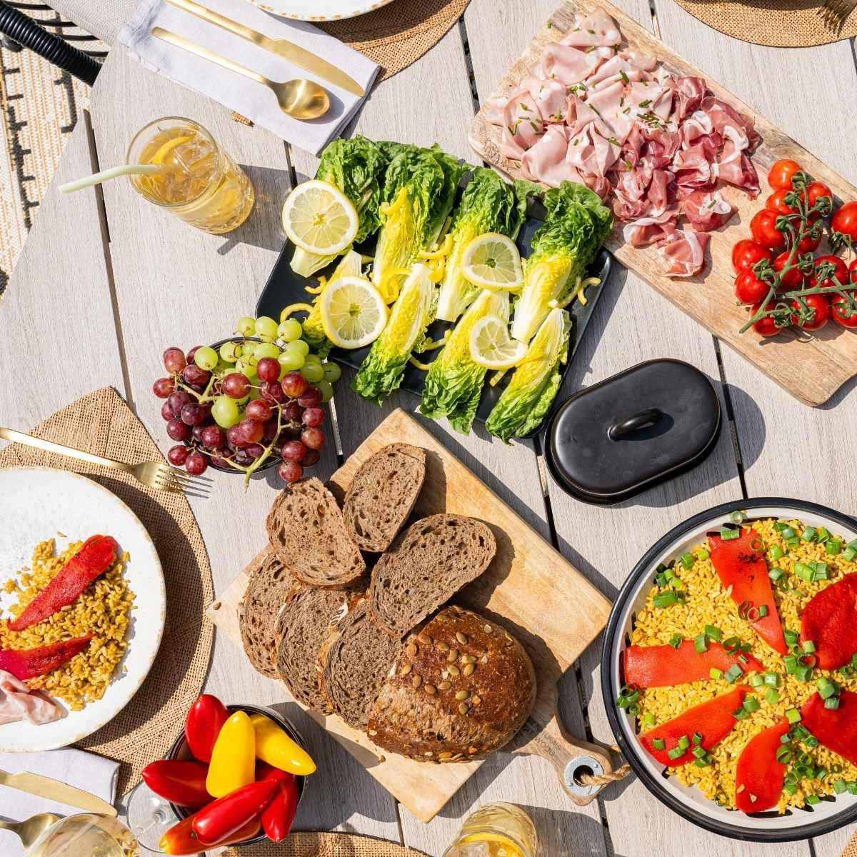 Table with food: bread, grapes, salad, tomatoes, ham, and other dishes. Outdoors, overhead view.