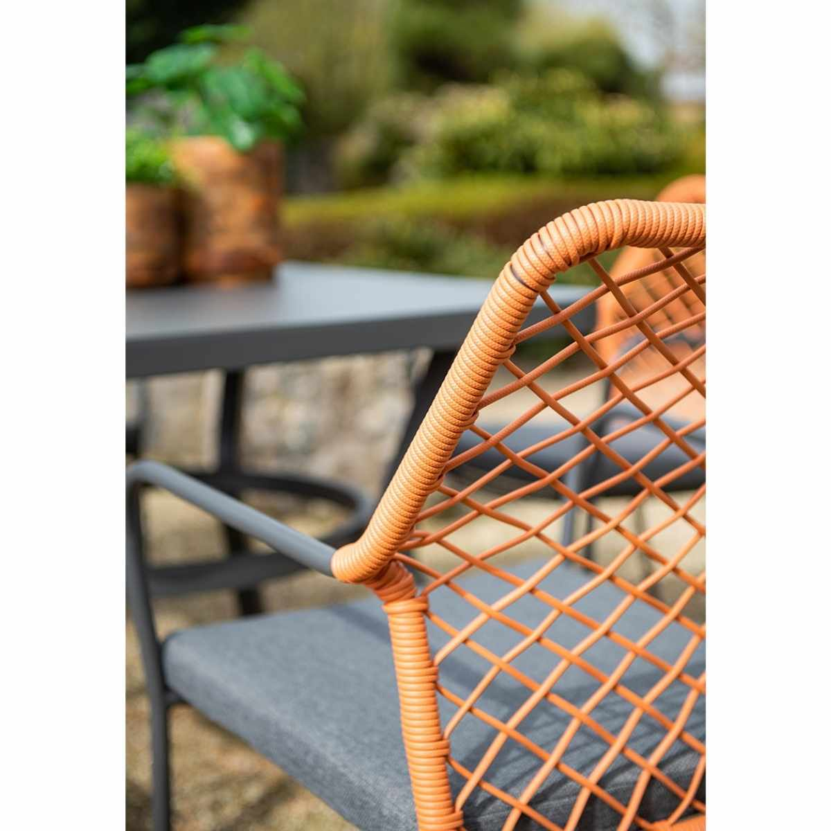 Close-up of chair. Orange rope design, gray cushion, and gray frame. Outdoors, blurred background.