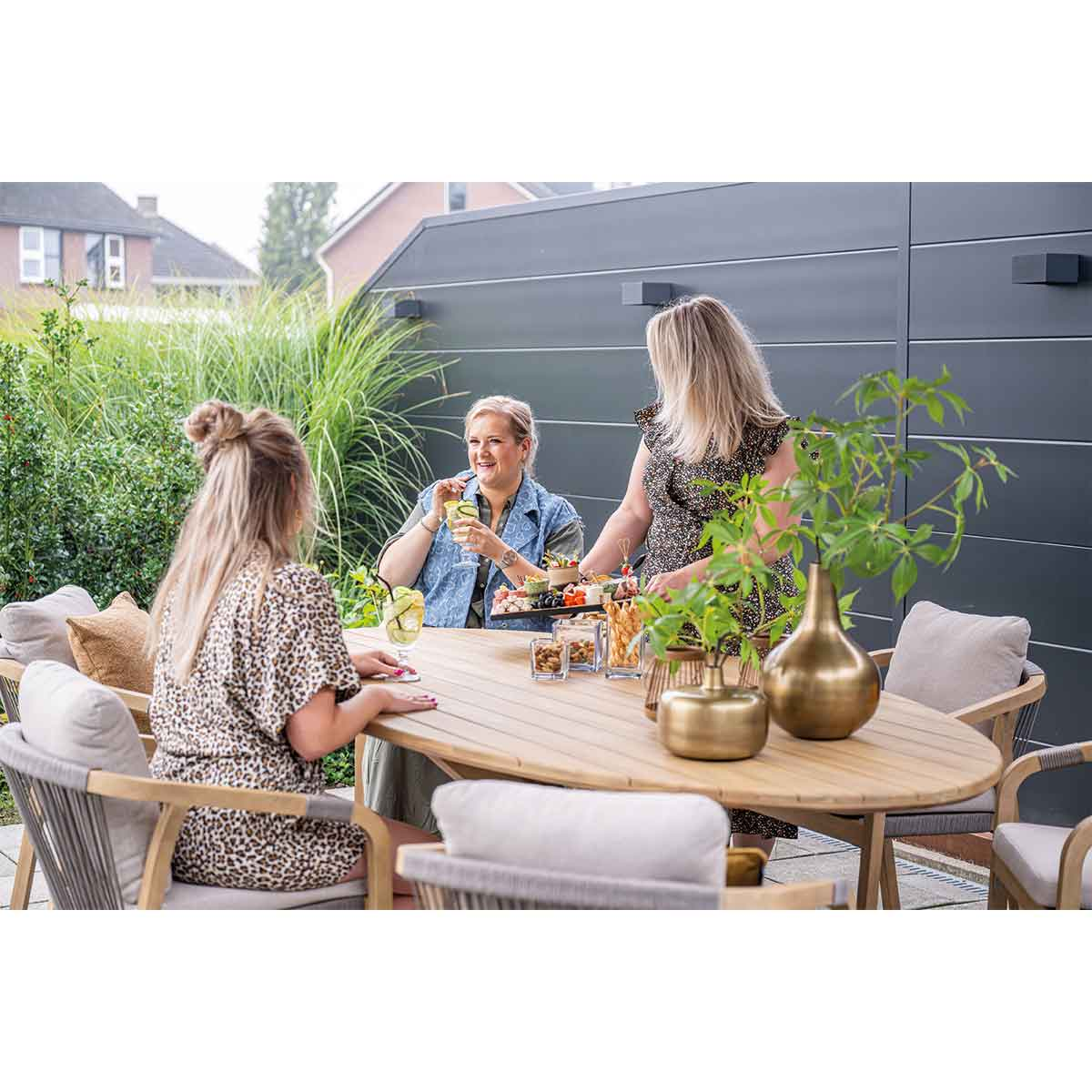 Drie mensen zitten rond een houten tafel met eten en drinken. Groene planten op de achtergrond.