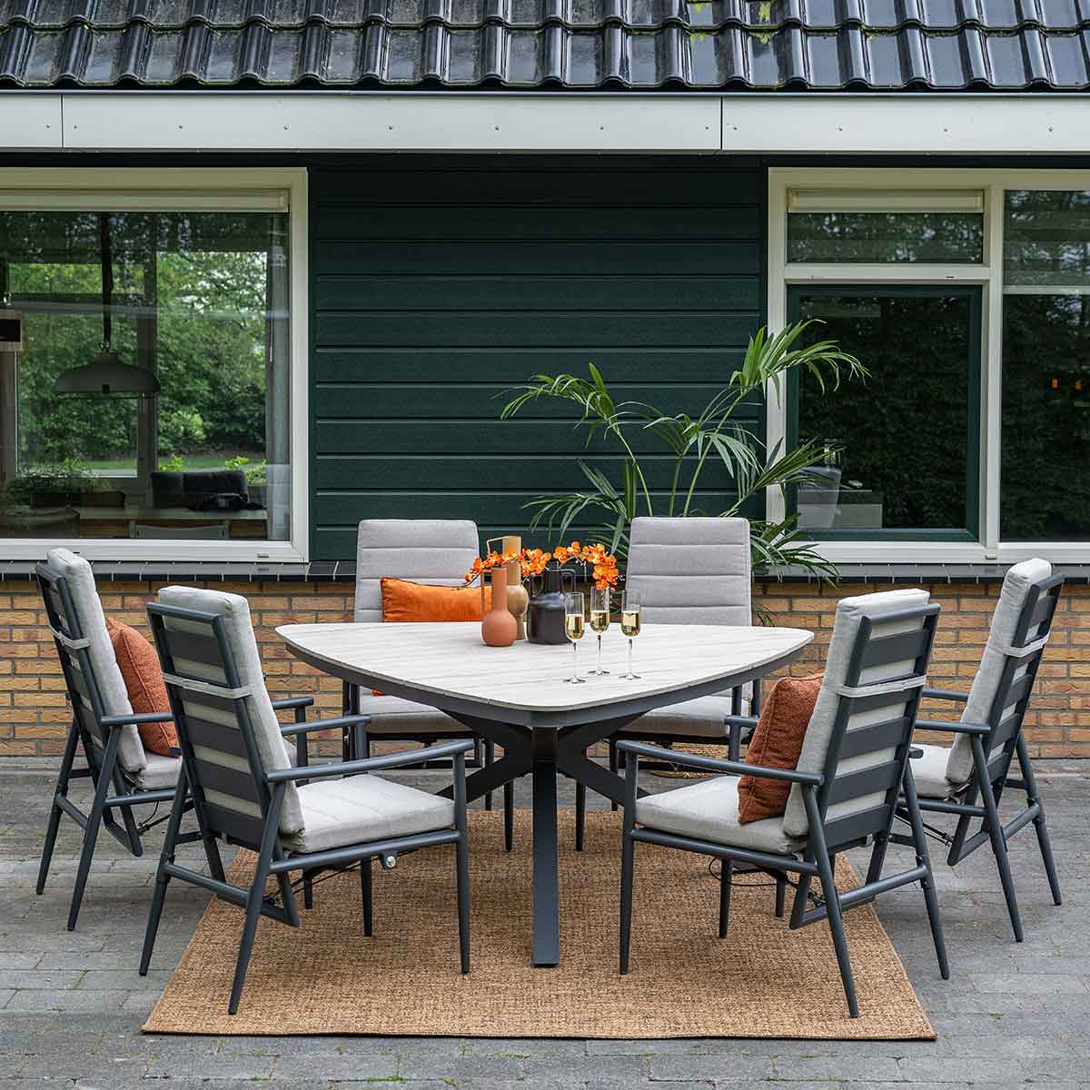 Outdoor dining set with a square table and six chairs. Green wall and brick ground are visible.