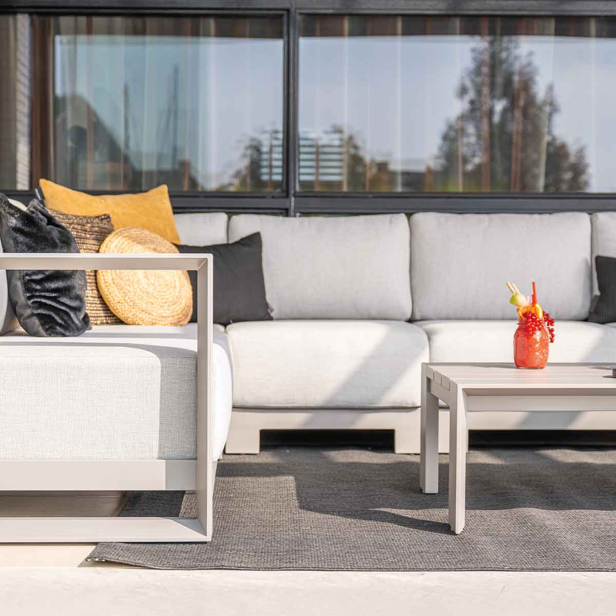 Outdoor scene with gray sofa, pillows, coffee table, and a drink. Windows in the background.