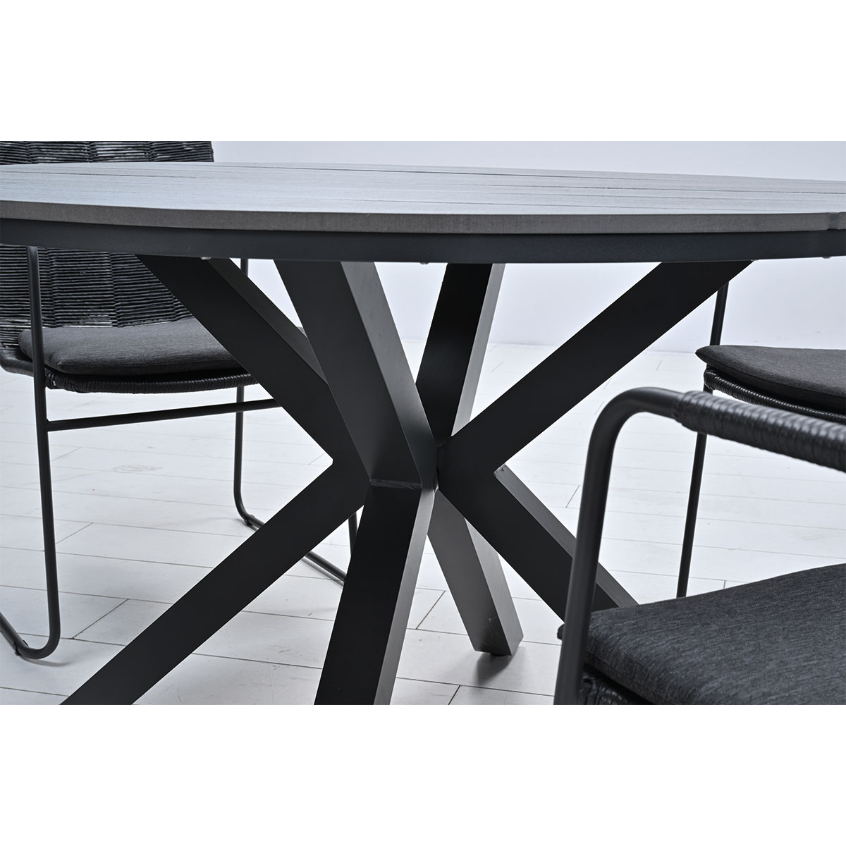 Close-up van een tafel met grijs blad en gekruiste zwarte poten. Twee stoelen zijn zichtbaar.