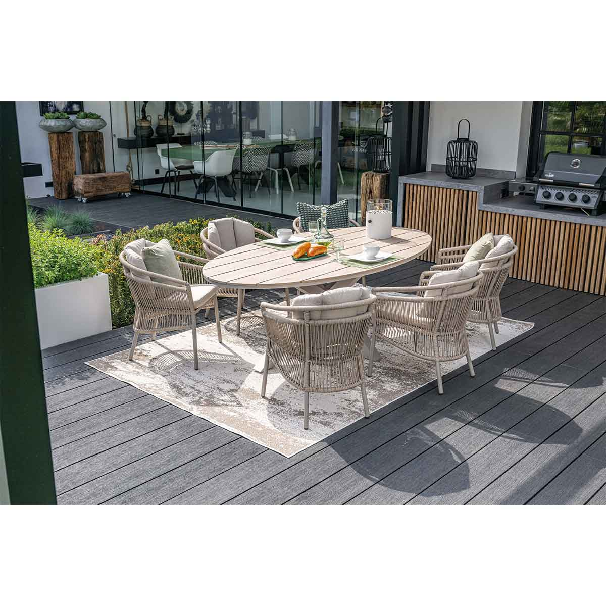 Outdoor dining set on a deck. Oval table with six chairs. Greenery in the background.
