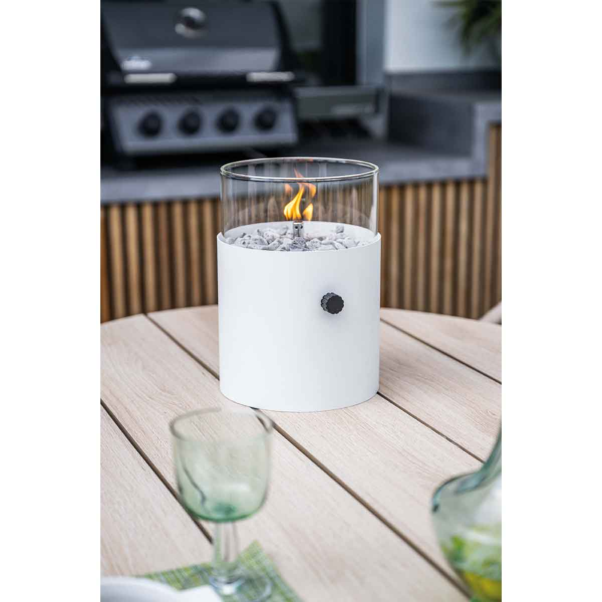 White outdoor fire pit with glass enclosure, rocks, and a flame. Set on a wooden table.