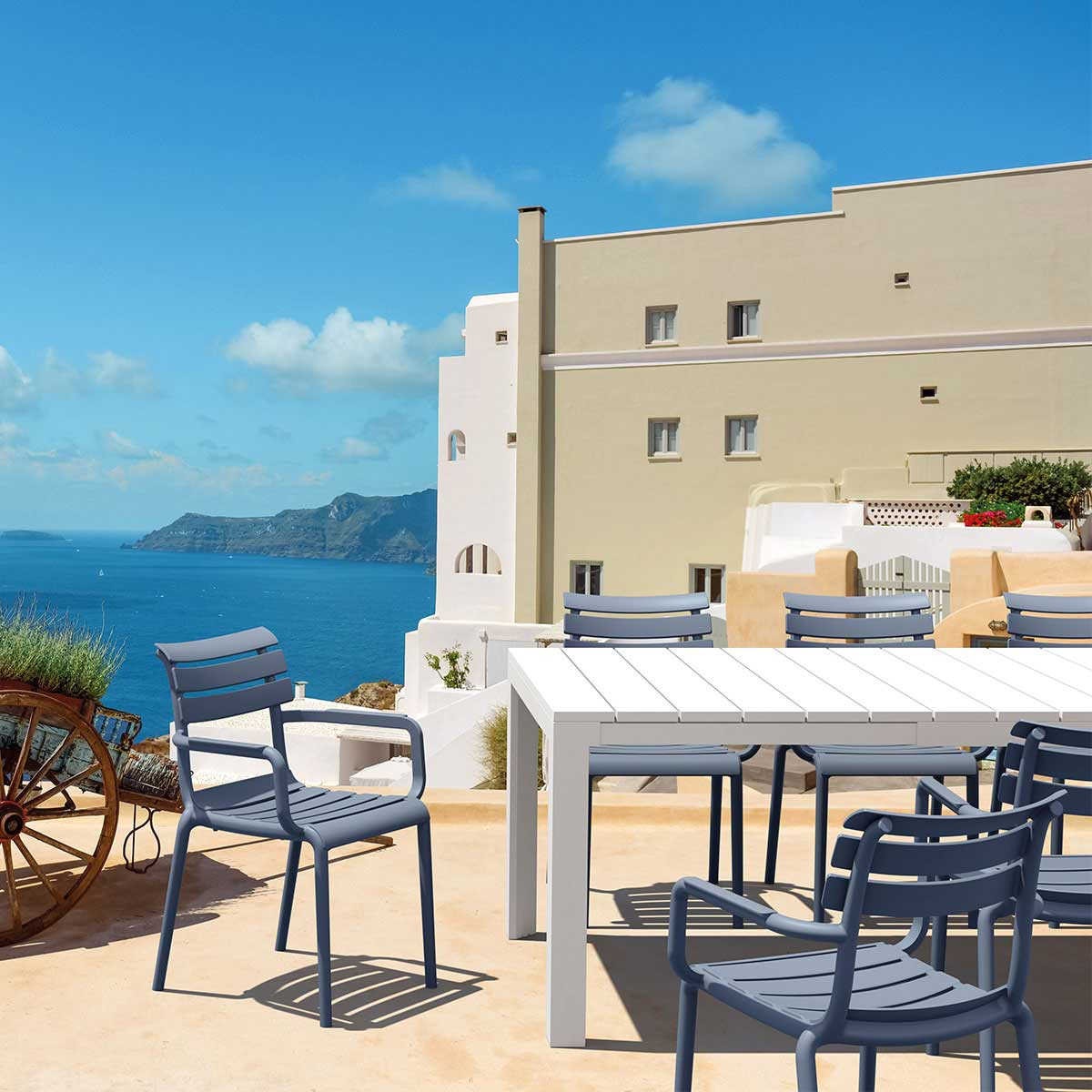 Scène extérieure avec chaises bleues, table blanche et bâtiments. Mer et ciel en arrière-plan.