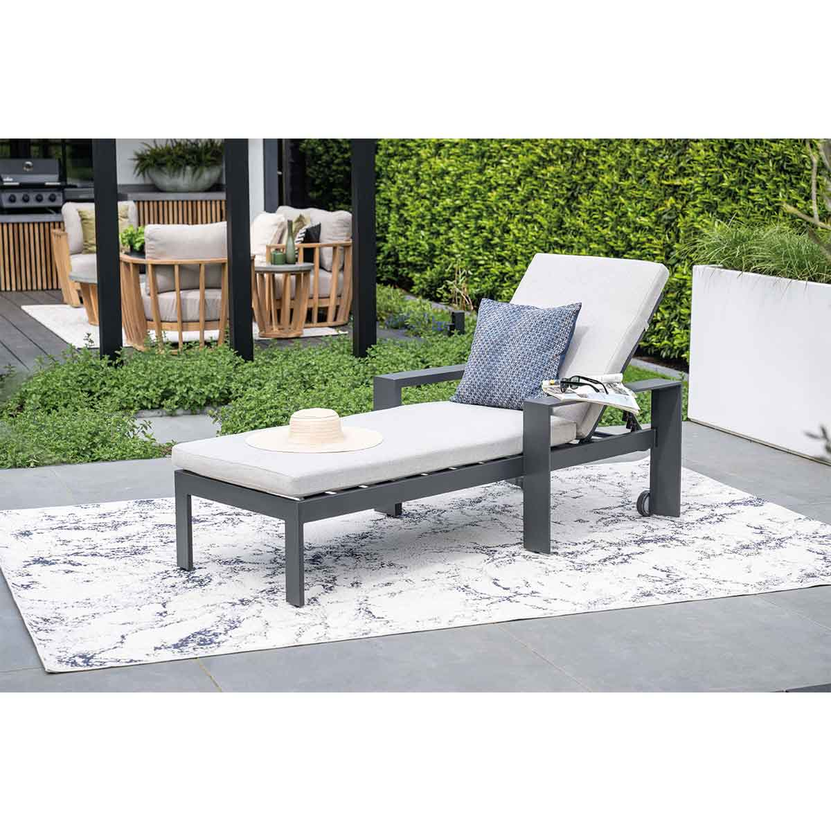 Gray chaise lounge on a patterned rug, hat, pillow, and magazine nearby.