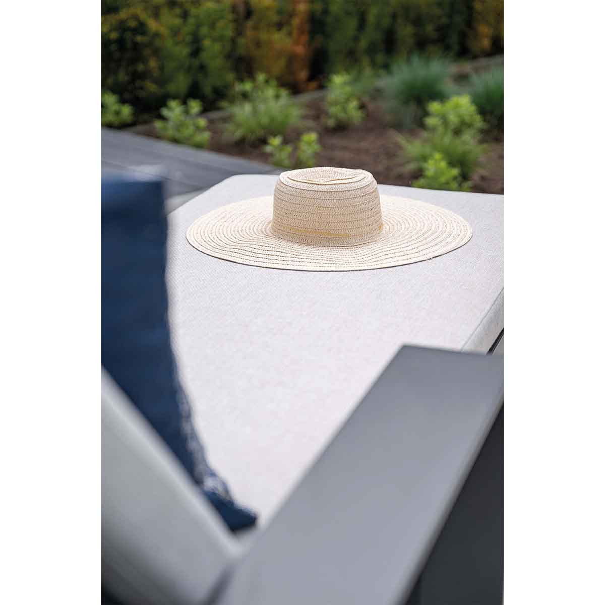A straw hat sits on a gray outdoor chaise lounge, green plants in the background.