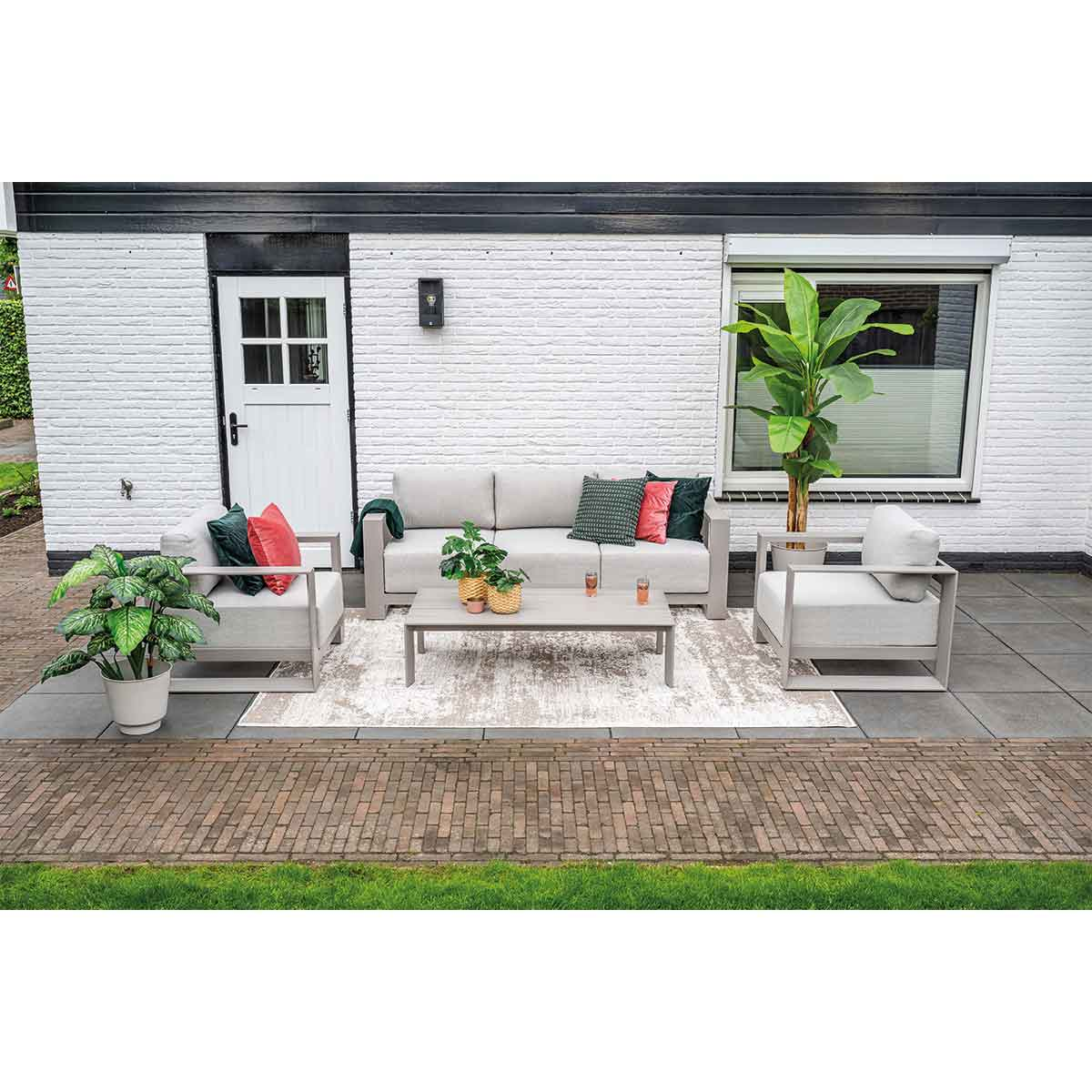 Coin salon extérieur avec canapé, deux chaises et table basse. Patio en briques, mur blanc et plantes.