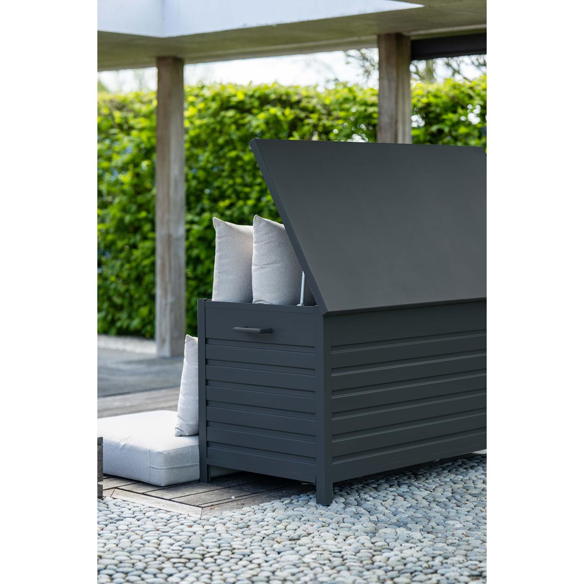 Un coffre de rangement gris foncé avec couvercle ouvert, coussins à l'intérieur et jardin en arrière-plan.