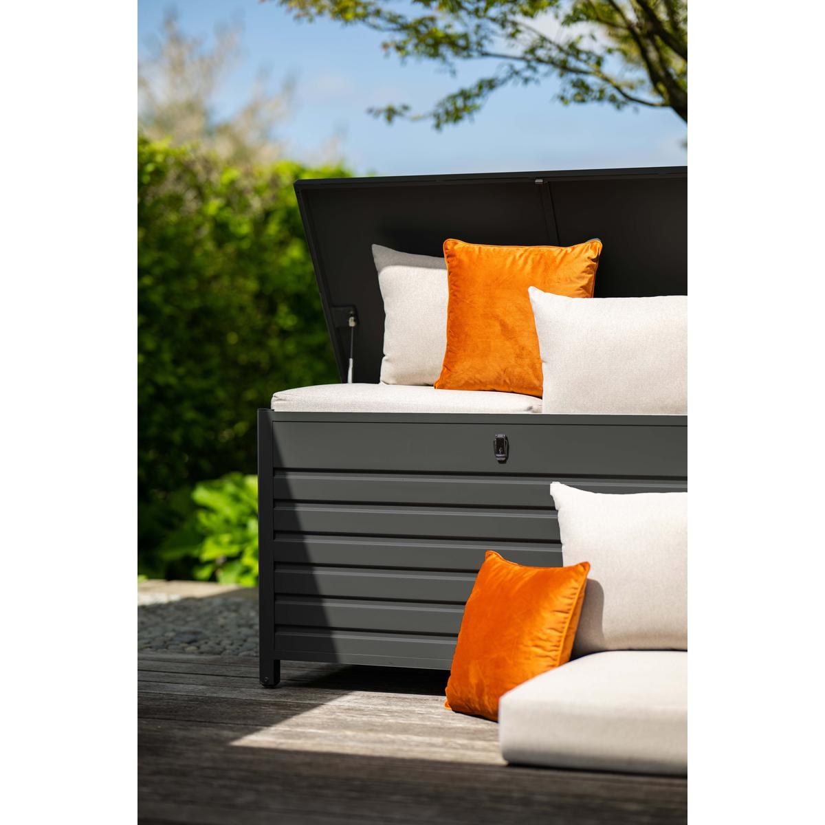 Un coffre de rangement gris foncé avec couvercle ouvert, coussins orange et blancs. Fond de jardin.