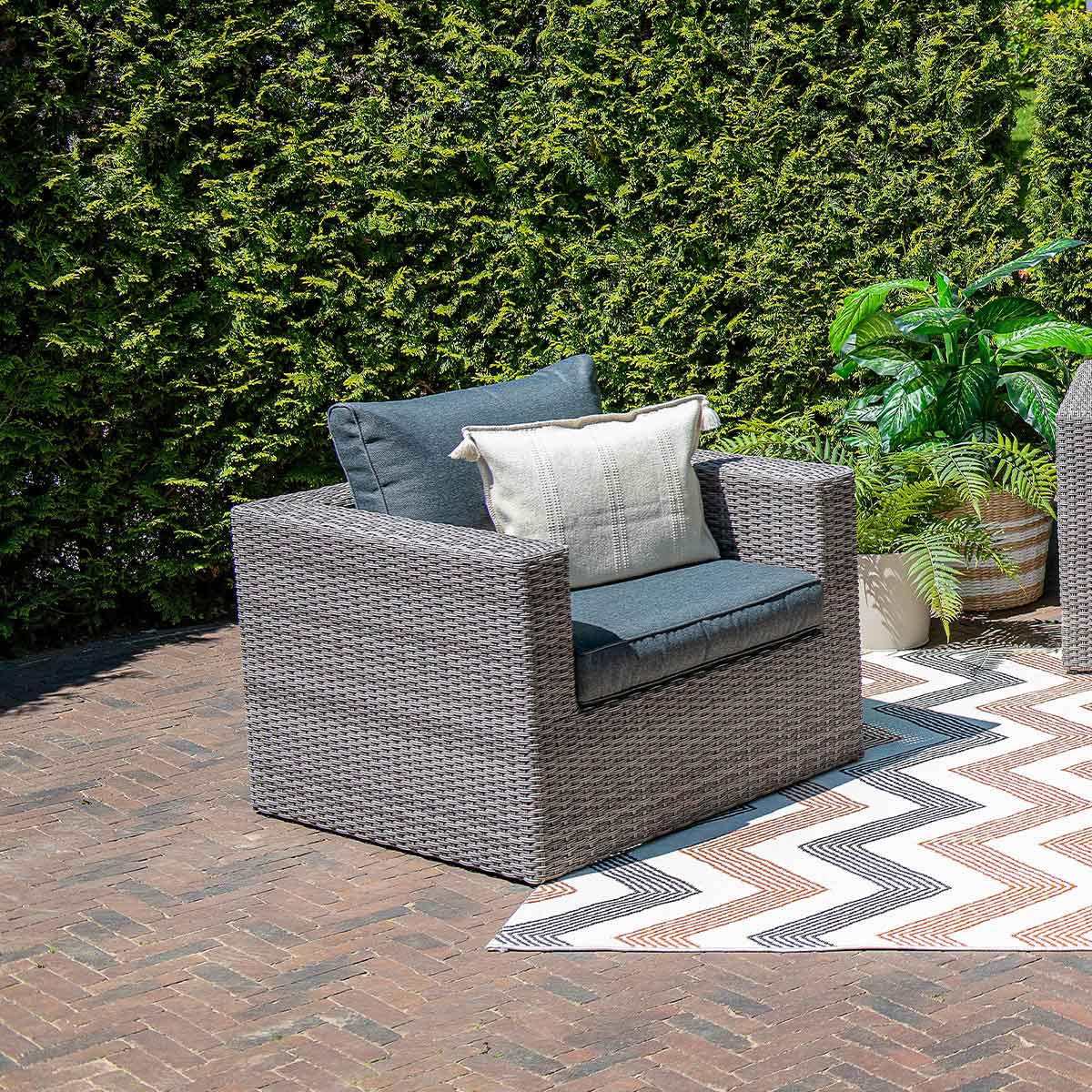 Grey wicker chair with a cushion and pillow on a patio with brick and rug, green hedge in the background.