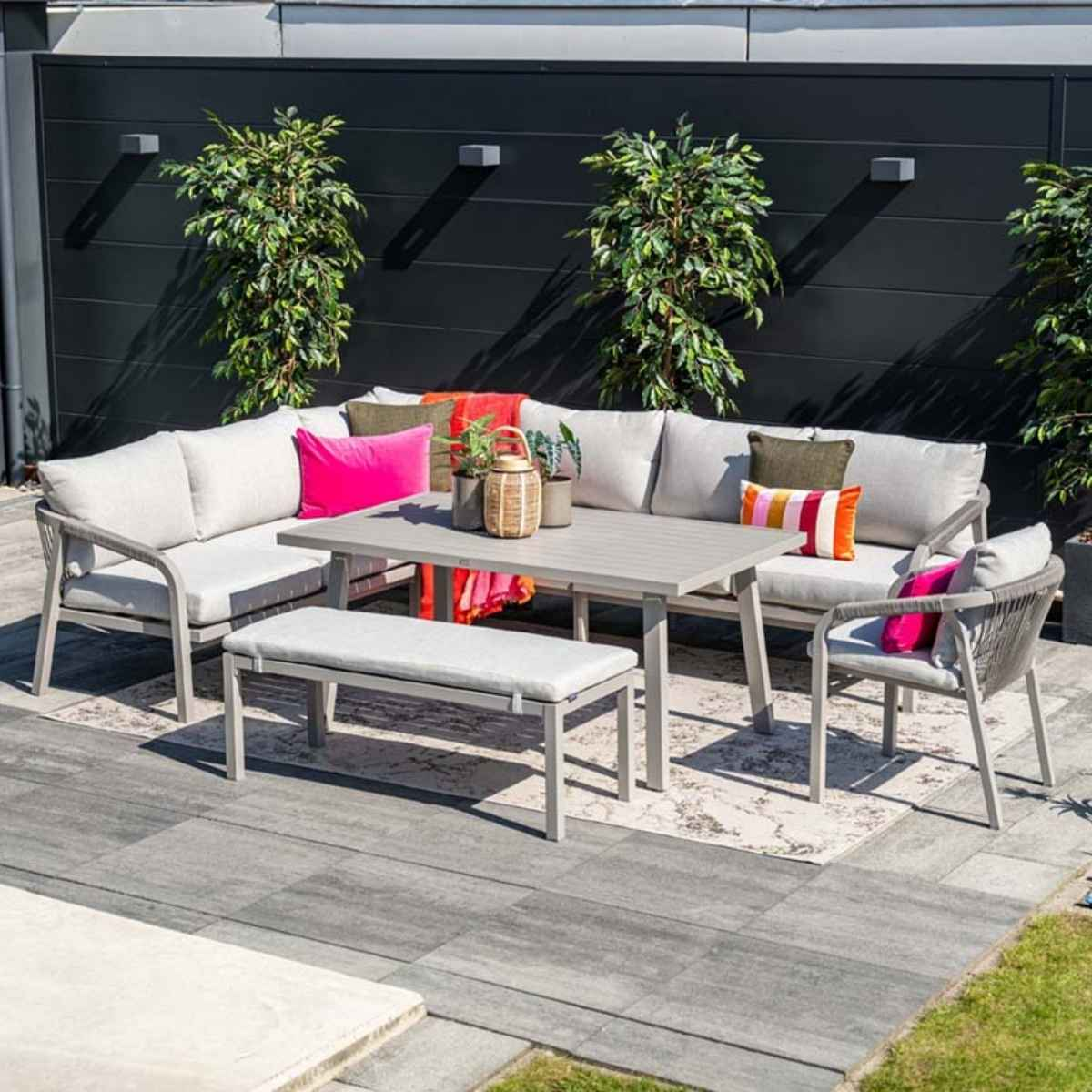 Ensemble de sièges d'extérieur avec table, banc, chaises et coussins gris. Plantes vertes et mur sombre en arrière-plan.