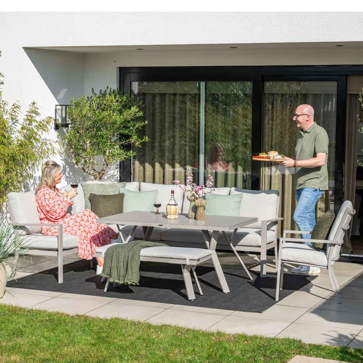 Scène de patio extérieur. Personnes, canapé gris, table et chaises. Nourriture, vin et plantes présents.