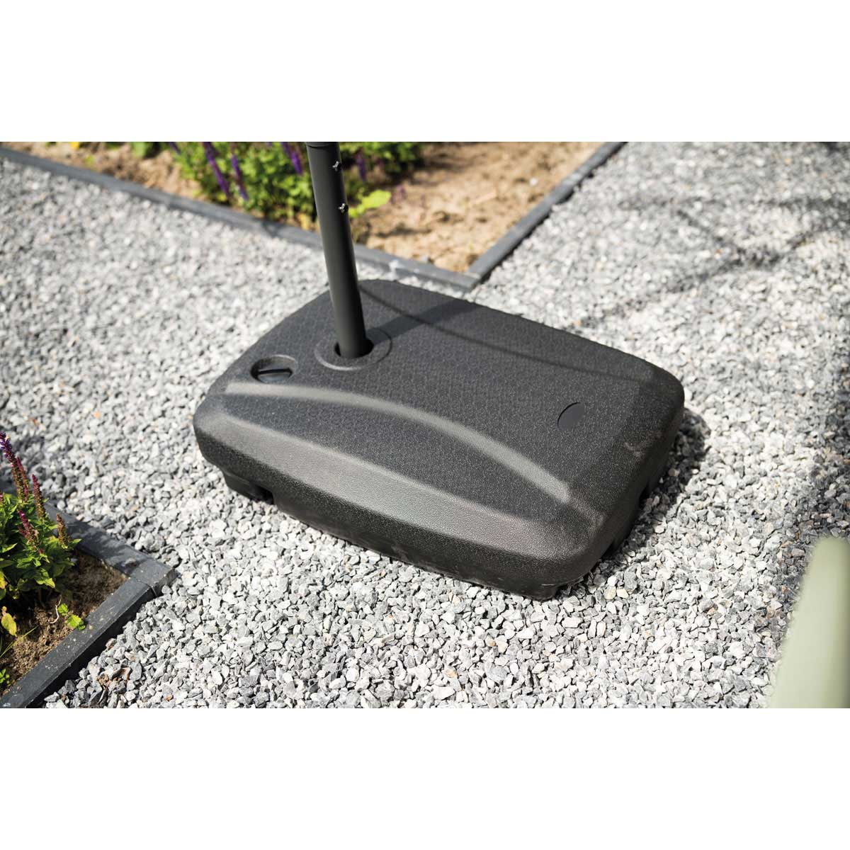 A black umbrella base on a gravel surface. The base is rectangular with curved sides.