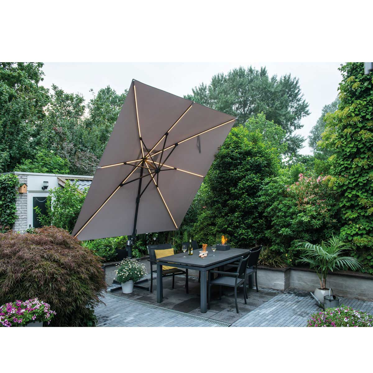 Scène de patio avec un grand parasol incliné au-dessus d'une table et des chaises, entouré de verdure.
