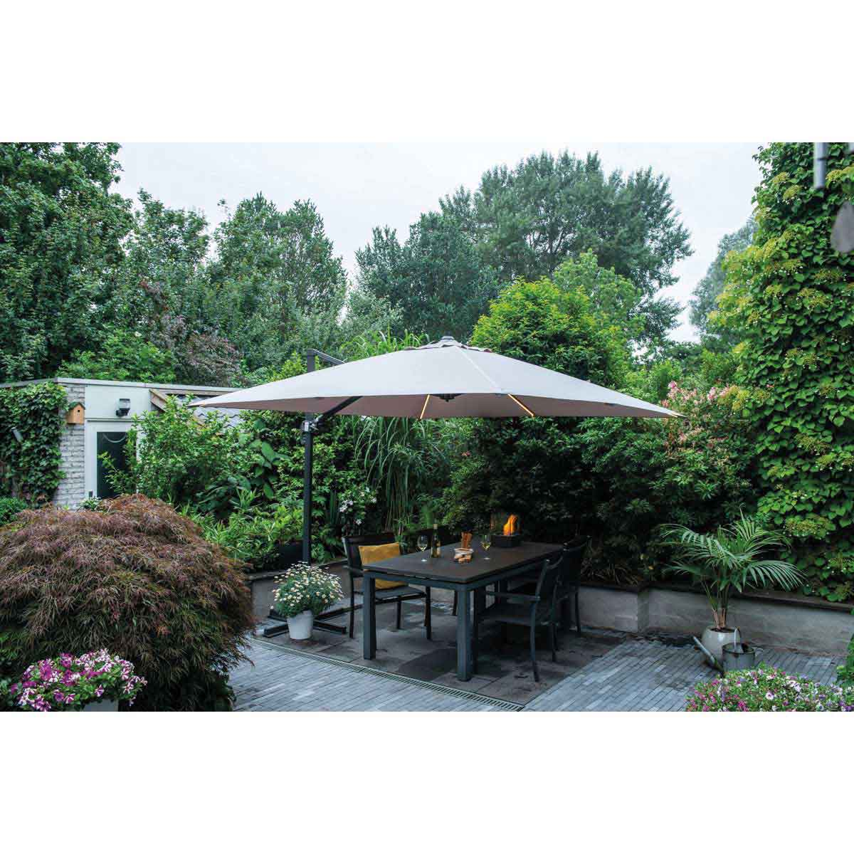 Scène de patio avec un grand parasol incliné au-dessus d'une table et des chaises, entouré de verdure.