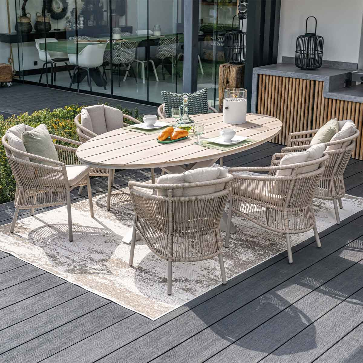 An outdoor dining set on a wooden deck with a woven rug. The set includes a table and chairs. The setting is sunny.