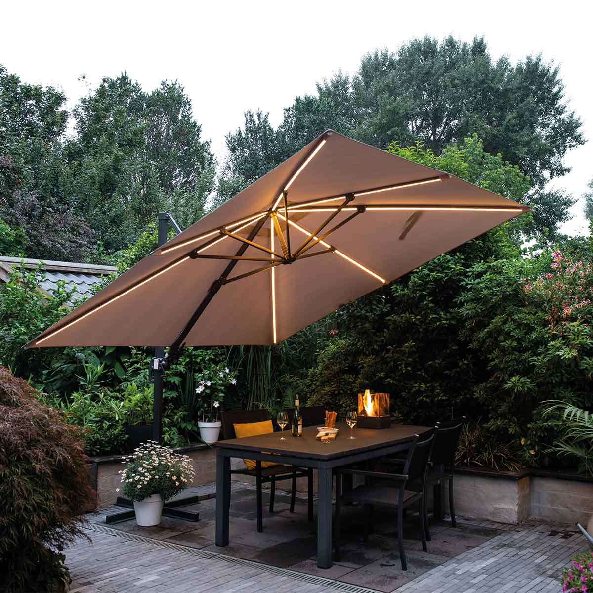 Un parasol carré éclairé au-dessus d'une table dressée pour un repas, dans un jardin au crépuscule.