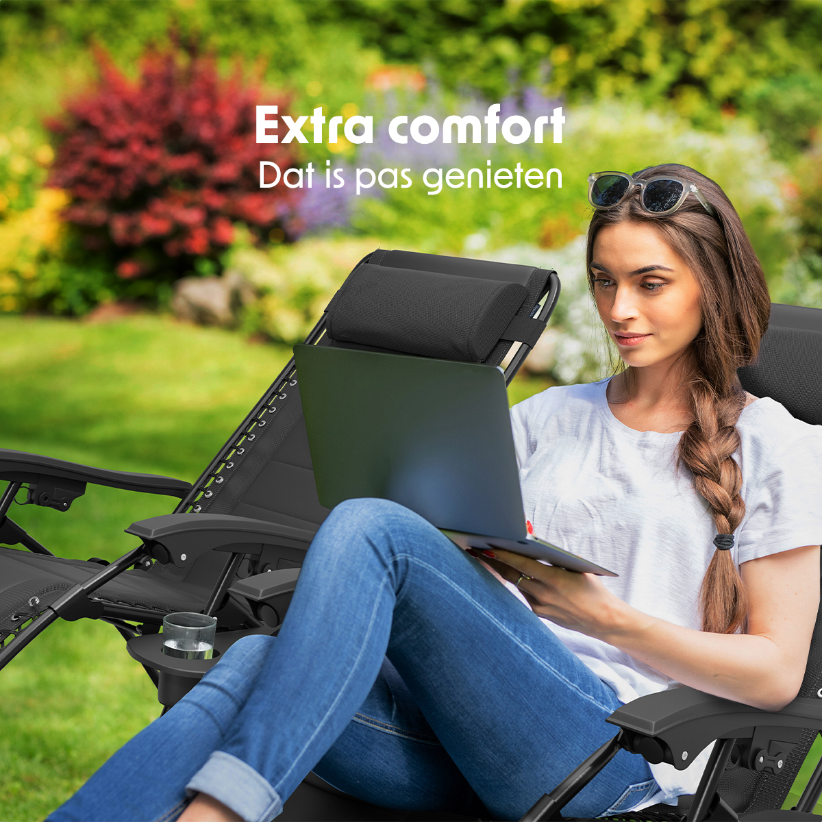 Een vrouw gebruikt een laptop terwijl ze in een zwarte ligstoel in een tuin zit. Zonnebril op haar hoofd.