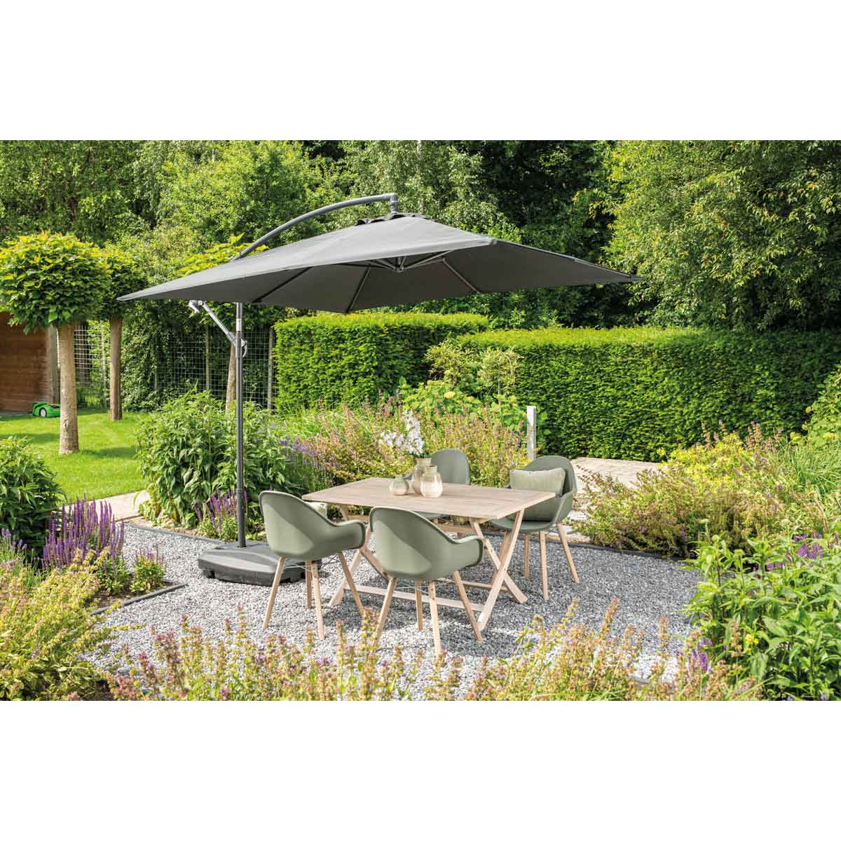Outdoor dining set: table, chairs, and a cantilever umbrella. Green bushes and gravel.