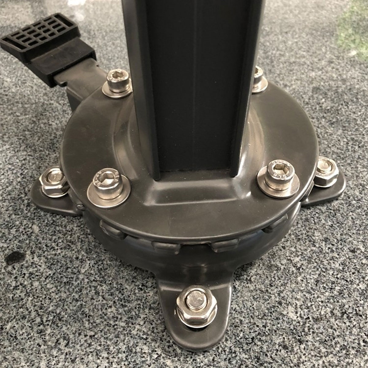 A close-up of a dark gray umbrella base with bolts on a stone surface. A few small water spots.